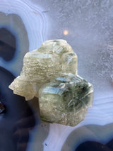 Translucent pale green fluorapatite crystal cluster with defined hexagonal shapes, resting on a smooth banded agate surface with blue and white rings.
