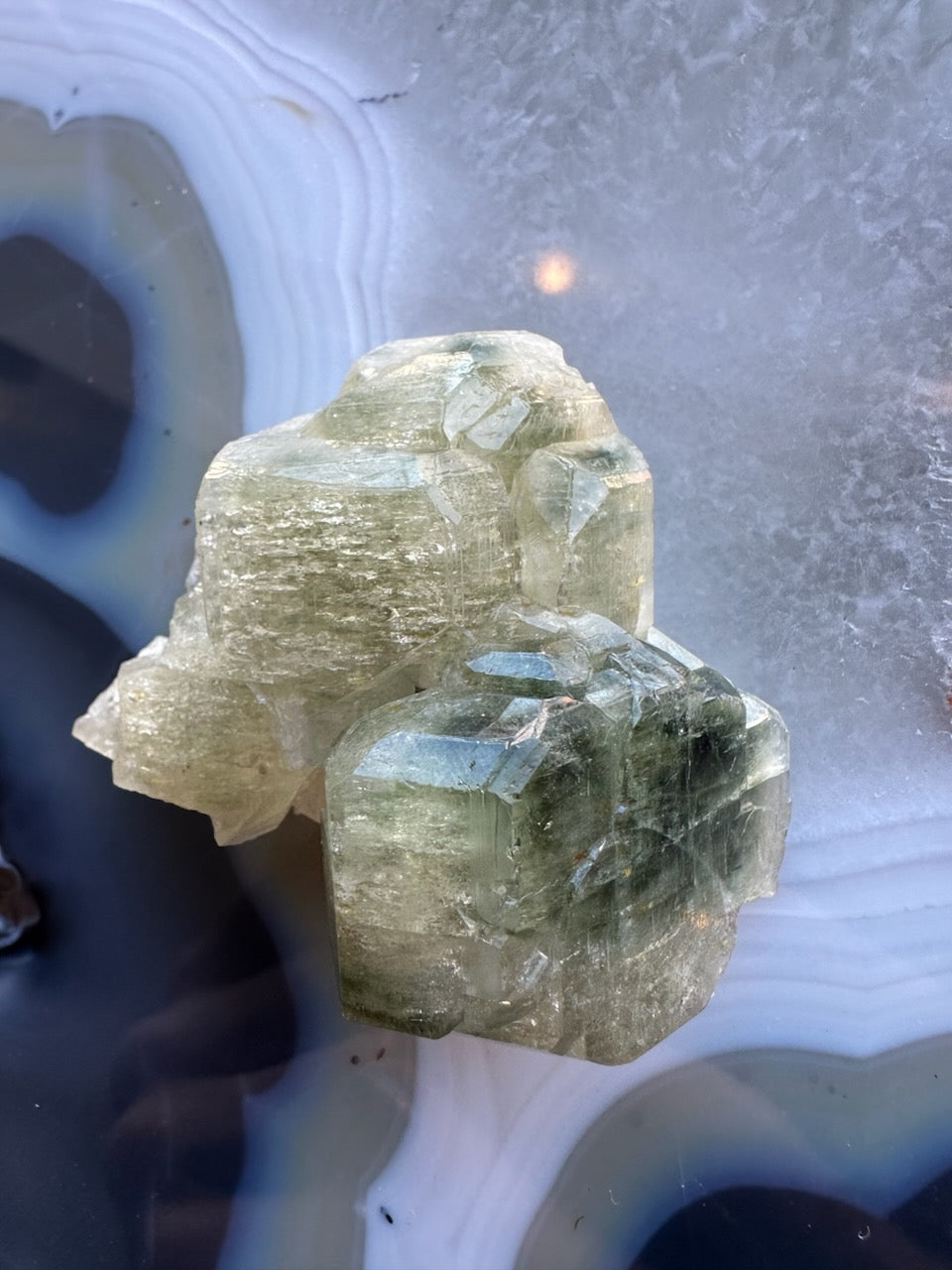 Translucent pale green fluorapatite crystal cluster with defined hexagonal shapes, resting on a smooth banded agate surface with blue and white rings.