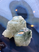 Two translucent pale green fluorapatite crystal cluster pieces resting on a polished blue agate surface with concentric light and dark bands and soft reflections.