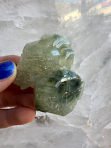 Hand holding a translucent pale green fluorapatite cluster with angular crystal formations against a white crystalline background, showing multiple faceted surfaces.