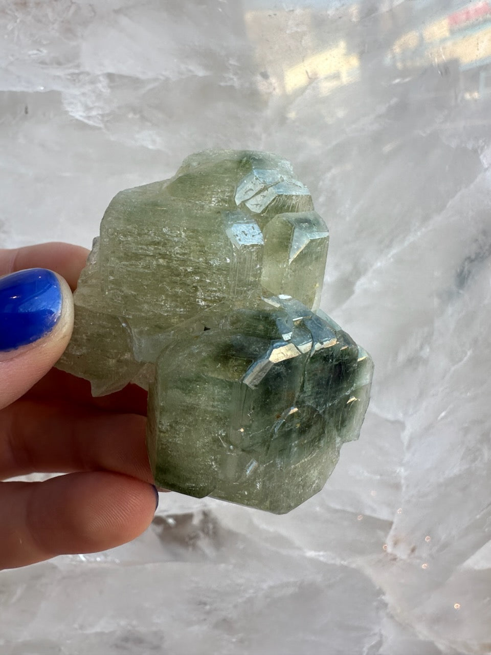 Hand holding a translucent pale green fluorapatite cluster with angular crystal formations against a white crystalline background, showing multiple faceted surfaces.
