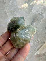 Hand holding a translucent pale green fluorapatite crystal cluster with defined angular faces against a blurred light gray crystalline background.