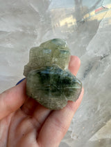 Hand holding a translucent green and gray fluorapatite crystal cluster against a light, textured background, highlighting its layered, blocky structure.