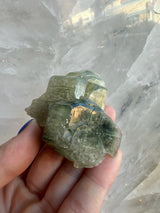 Greenish translucent fluorapatite crystal cluster held between fingers, showing hexagonal facets and glossy surfaces against a light background.