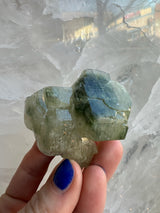 Hand holding a translucent green fluorapatite crystal cluster with visible internal fractures against a background of larger white crystalline formations.
