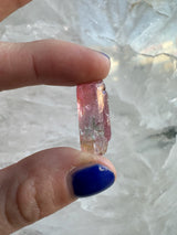 Close-up of a hand with blue nail polish holding a transparent pink and green watermelon tourmaline crystal against a pale, textured background.