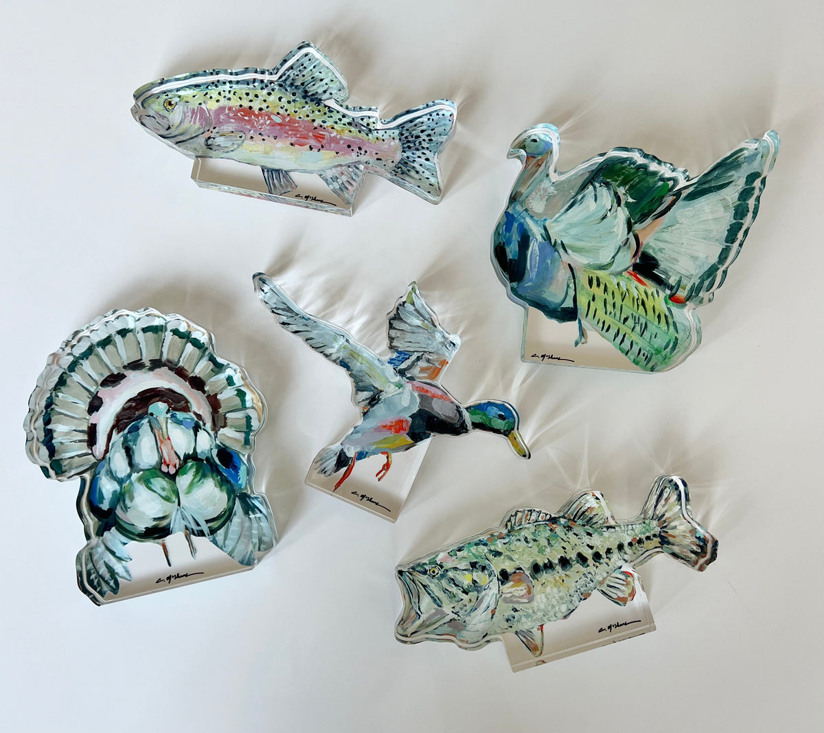 Five painted acrylic tabletop art pieces featuring a turkey with fanned tail, a flying mallard duck, a green-winged teal in flight, a rainbow trout, and a bass with speckled markings, each mounted on clear stands.