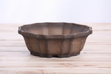 Brown ceramic bonsai pot with a flower-shaped rim and textured surface, sitting on a light wooden slatted surface against a pale background.