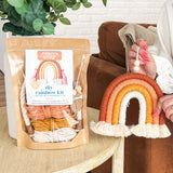Hand holding a warm-toned macrame rainbow with fringe, next to a packaged DIY rainbow kit with cotton cords in similar earth tones on a wooden table.