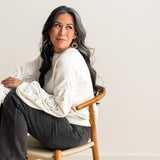Woman seated sideways on wooden chair wearing macrame knotted earrings and a white long-sleeve top with rose embellishments on the sleeves.