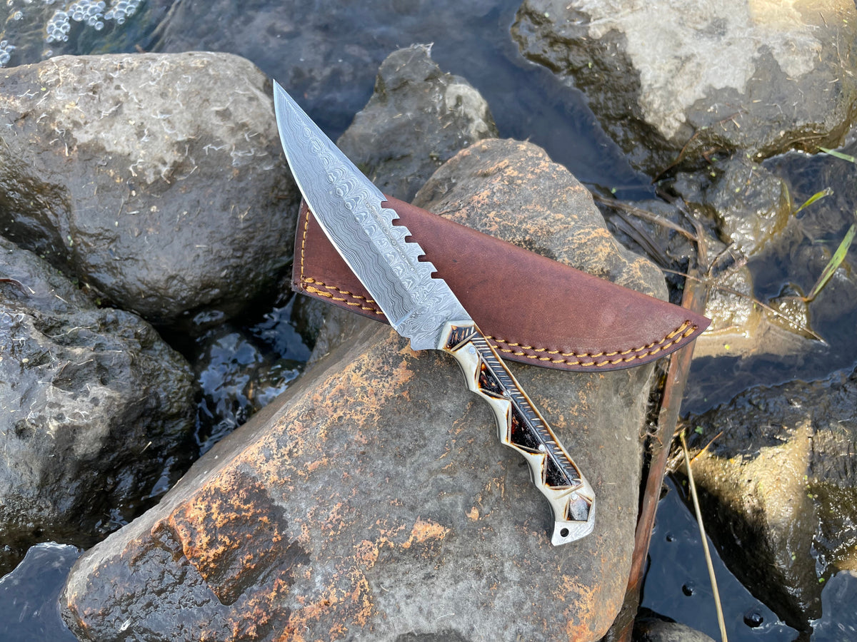 Handmade High Carbon Damascus Steel Hunting Knife with burnt bone handle and leather sheath laid on a wood surface