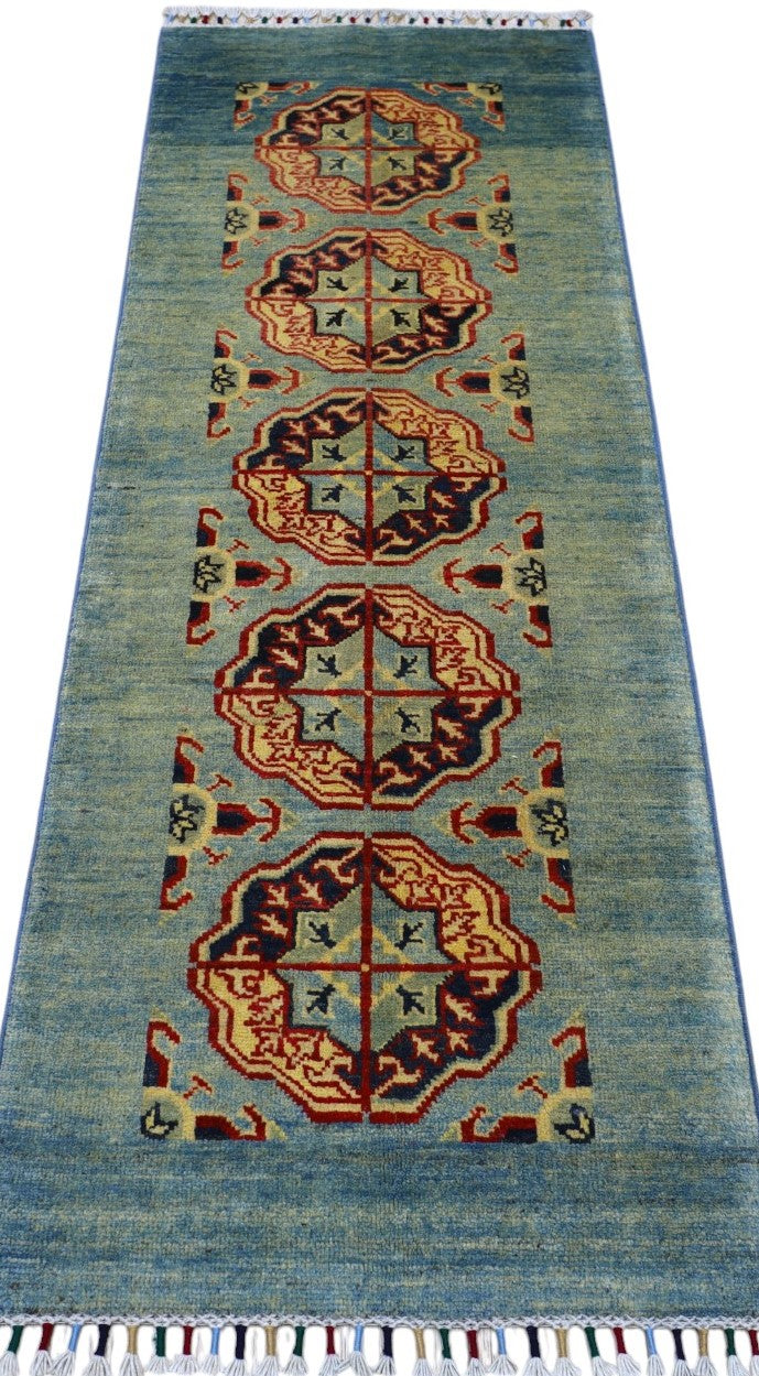 Long rectangular Afghani Kazak wool rug with a muted blue-green field and repeating hexagonal medallions in cream, red, brown, and black, finished with white fringe.