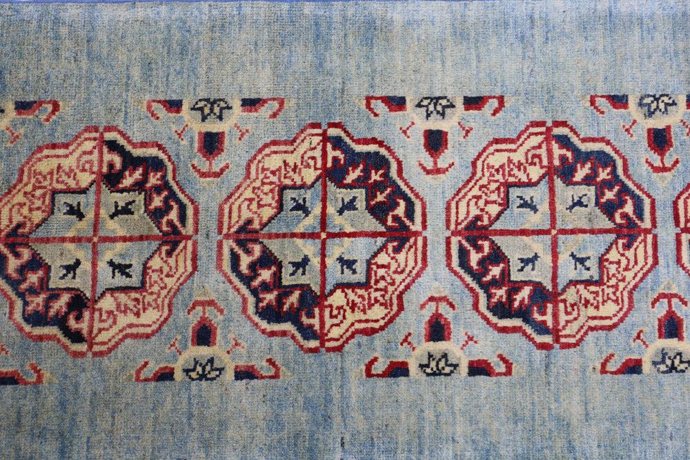 Section of a hand-knotted Afghani Kazak wool rug with repeating octagonal medallions in red, dark blue, and ivory on a light blue field, featuring geometric motifs.