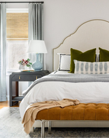Bed with beige curved headboard, white bedding, green velvet pillows, blue-gray patterned lumbar pillow, and a tan tufted bench at foot in a softly-lit bedroom.