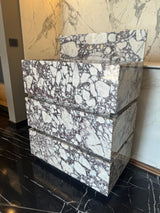 Freestanding Calacatta Viola marble vanity with bold white and purple veining, three horizontal drawers stacked in a sculptural cubic form on a black marble floor.