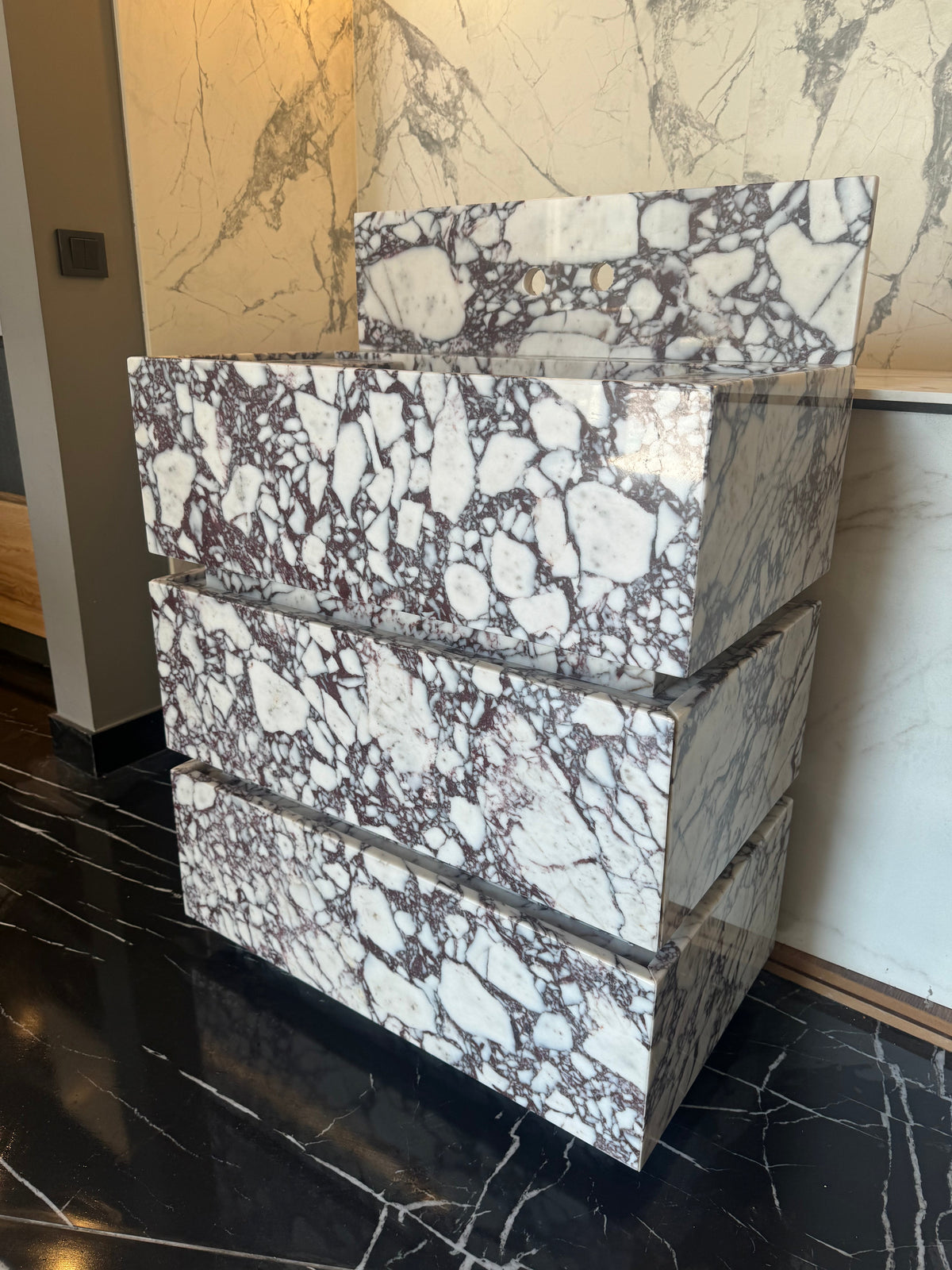 Freestanding Calacatta Viola marble vanity with bold white and purple veining, three horizontal drawers stacked in a sculptural cubic form on a black marble floor.