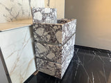 Freestanding Calacatta Viola marble vanity with three stacked rectangular drawers featuring white and dark purple veining, positioned next to a marble wall and black floor tiles.