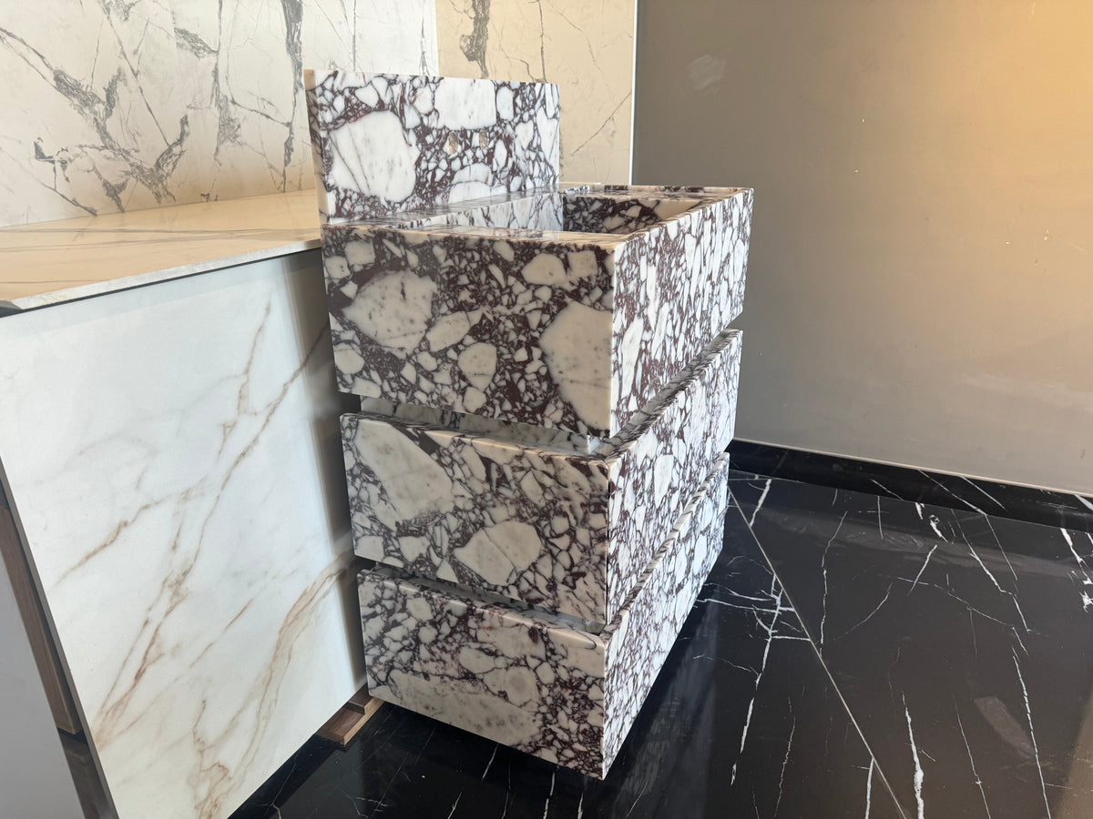 Freestanding Calacatta Viola marble vanity with three stacked rectangular drawers featuring white and dark purple veining, positioned next to a marble wall and black floor tiles.