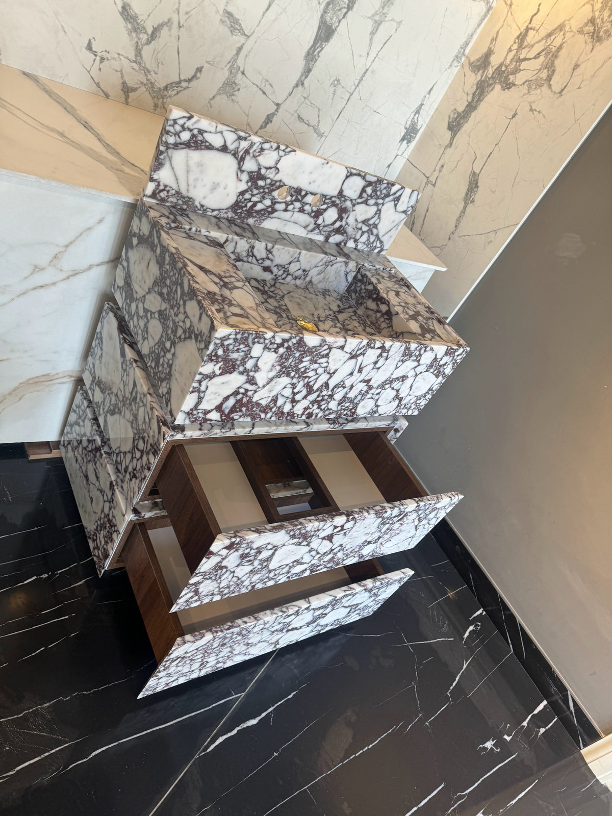 Calacatta Viola marble freestanding vanity with an integrated sink and two open drawers, featuring deep purple and brown veining on a white base, set against matching marble walls and a black floor.