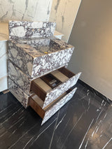 Freestanding marble vanity with Calacatta Viola pattern featuring two open drawers revealing wooden interiors, set against a marble wall and black marble floor with white veining.