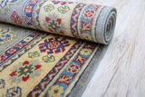 Partially rolled Afghani Kazak wool rug with beige background, featuring red, green, blue, and yellow floral and geometric patterns, displayed on light wood floor.