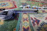 Close-up of a hand-knotted Afghani Kazak wool rug with colorful geometric and floral patterns, showing part of a measuring tape extended across its woven fringe.