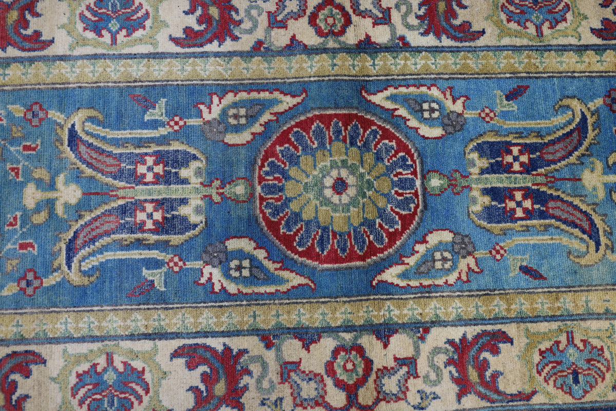 Section of a hand-knotted Afghani Kazak wool rug featuring a central red and green floral medallion on a bright blue background with surrounding intricate floral motifs in cream and red.