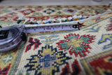Close-up of a hand-knotted Afghani Kazak wool rug with intricate geometric patterns in red, blue, yellow, and green, alongside a tape measure showing precise detail.