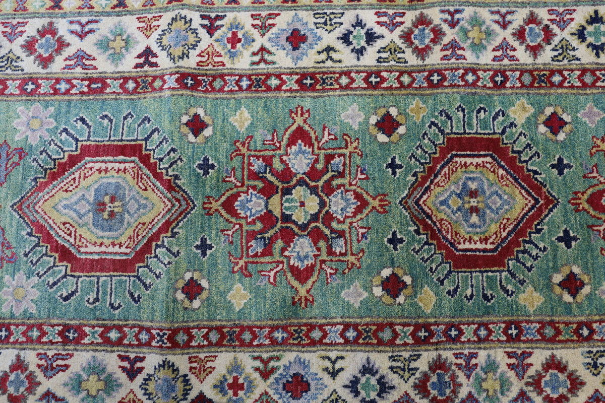 Section of a hand-knotted Afghani Kazak wool rug with a pale green background, featuring geometric medallions in red, blue, and cream framed by floral motifs and multiple borders.