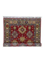 Hand-knotted Afghani Kazak wool rug with red background, geometric medallions in blue and beige, and intricate multicolored border and fringes, partially rolled at the bottom.
