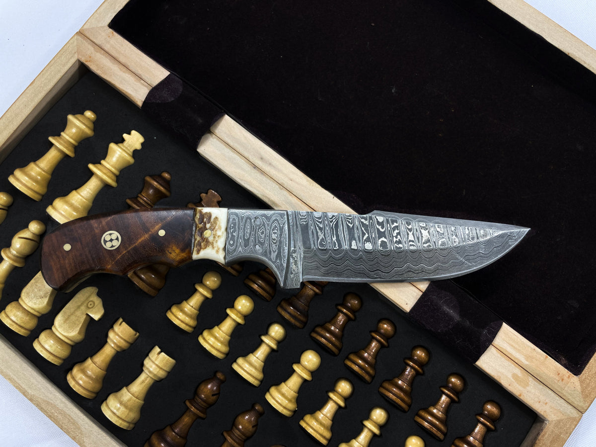 Damascus steel fixed blade hunting knife with intricate patterned blade and walnut handle with stag horn inlay, displayed inside an open wooden box on a chessboard background.
