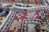 Close-up of a hand-knotted Afghani Kazak wool rug with red, blue, cream, and green geometric floral patterns and a measuring tape showing its thickness.
