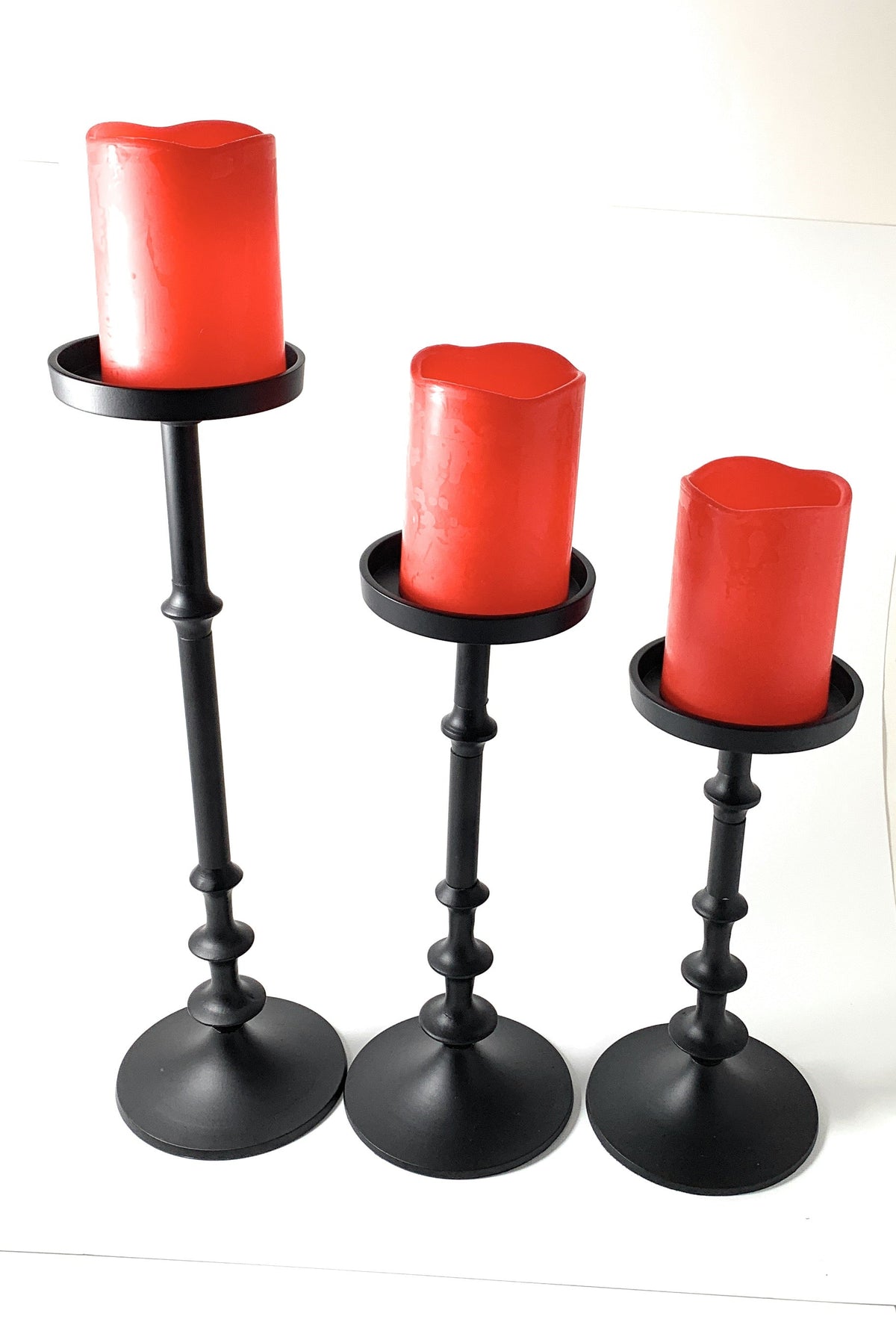 Set of three matte black metal candle holders with round bases, each holding a large red pillar candle of varying heights with melted tops.