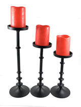 Set of three black metal candle holders of varying heights, each holding a red, worn-top pillar candle with melted edges.