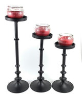 Set of three black metal candle holders of varying heights, each holding a red ribbed glass candle jar, arranged diagonally on a white background.
