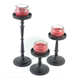 Set of three black metal pillar candle holders with turned stems, each holding a small red jar candle with vertical ridges, arranged by height on white background.