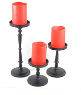 Set of three black metal pillar candle holders with turned detailing, each holding a red flameless candle of varying heights on a white background.