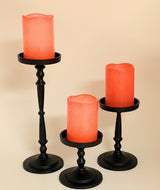 Set of three black metal pillar candle holders of varying heights, each holding an orange faux LED pillar candle with a slightly melted top edge.