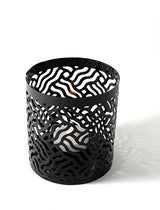 Black metal votive candle holder with an abstract wavy cutout pattern casting intricate shadows on a white surface.