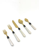 Flatware set with white pearlescent resin handles, antique-style black metal collar, and gold-toned stainless steel heads, including fork, knife, and spoons.