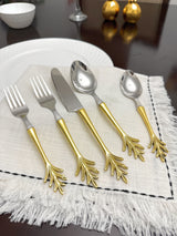 Stainless steel flatware set with golden handles shaped like delicate leaf branches, arranged on a textured off-white napkin on a wooden table.