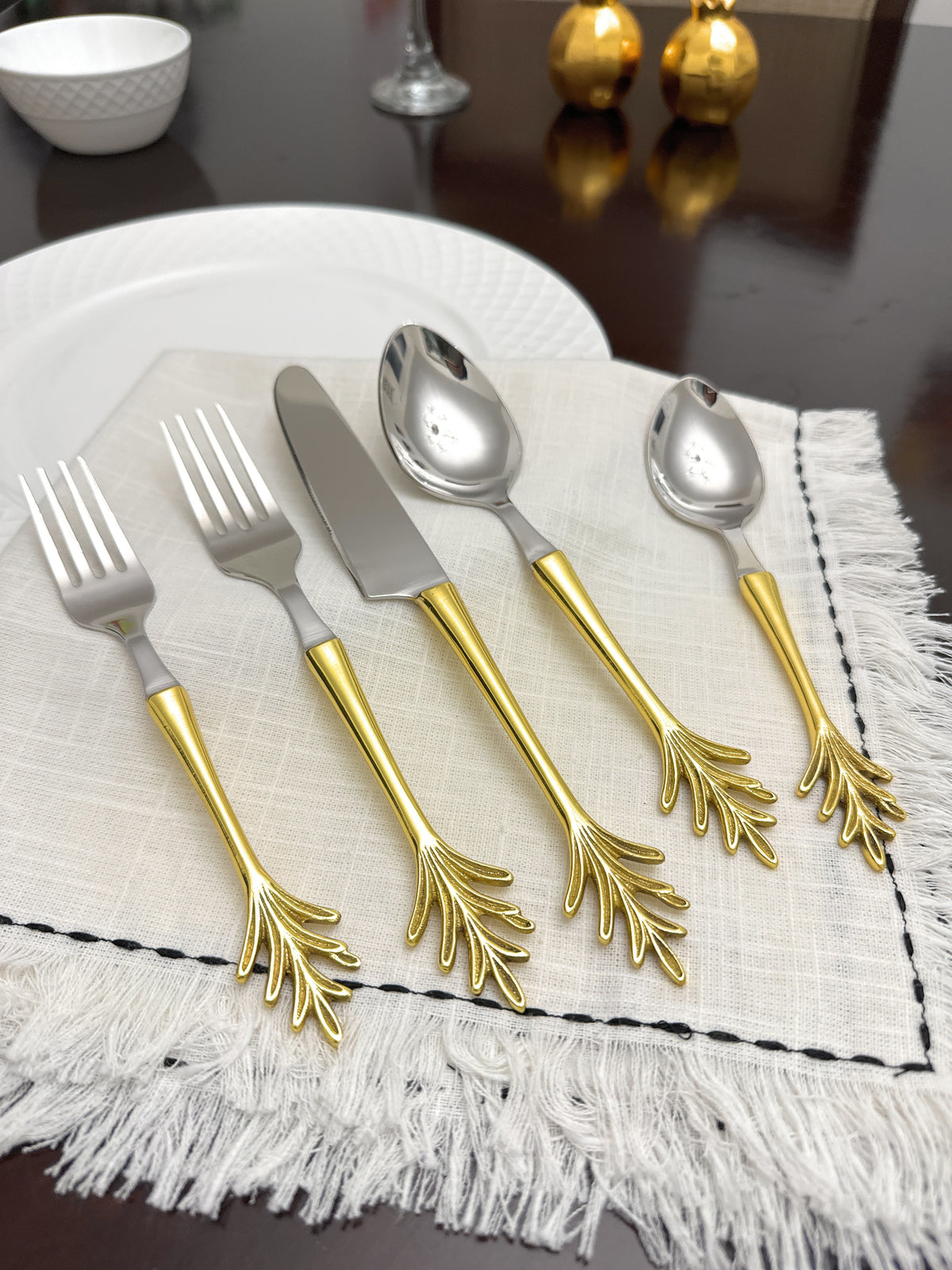 Stainless steel flatware set with golden handles shaped like delicate leaf branches, arranged on a textured off-white napkin on a wooden table.