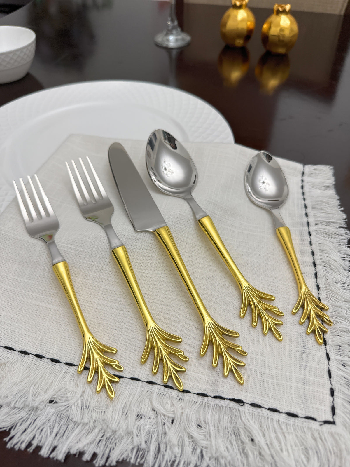 Five-piece flatware set with golden leaf-shaped handles and shiny silver utensil heads arranged on a fringed white cloth, placed on a dark wooden table.