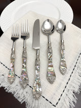 Stainless steel flatware set with ornate handles featuring iridescent abalone inlay and decorative silver-toned accents, displayed on a fringed white cloth.