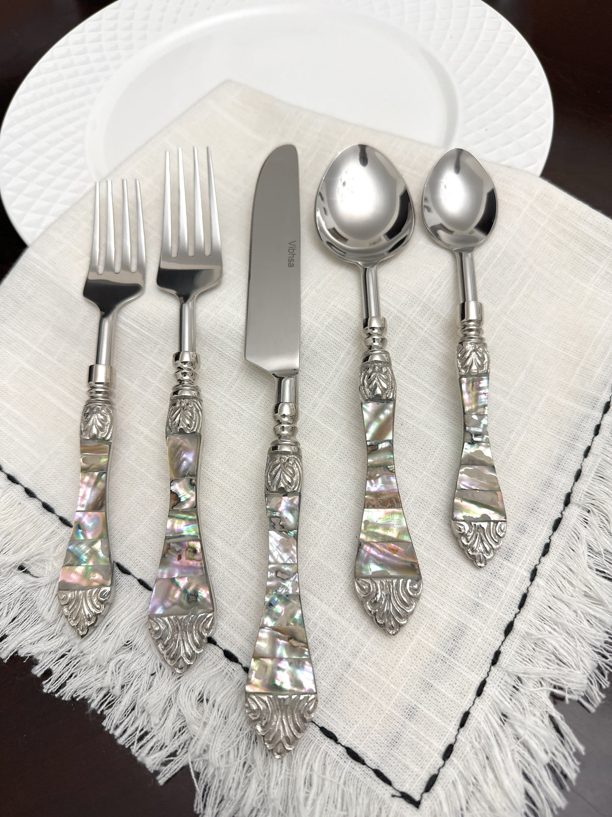 Stainless steel flatware set with ornate handles featuring iridescent abalone inlay and decorative silver-toned accents, displayed on a fringed white cloth.