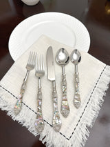 Five-piece stainless steel flatware set with handles featuring iridescent mother-of-pearl seashell patterns, arranged on a white fringed cloth atop a dark table near a white plate.