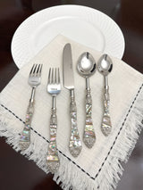 Five-piece flatware set with stainless steel utensils and iridescent seashell-patterned handles, arranged on a fringed beige napkin over a white textured plate.
