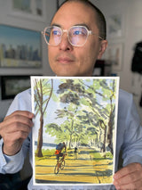 Person wearing glasses holds a watercolor painting of bikers riding along a tree-lined path by a river, with green and yellow foliage and blue sky visible.