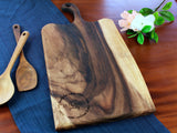 Rectangular live edge wooden cutting board with handle, showing rich dark and light wood grain patterns, placed on a blue cloth with wooden spoons and a small flower arrangement.