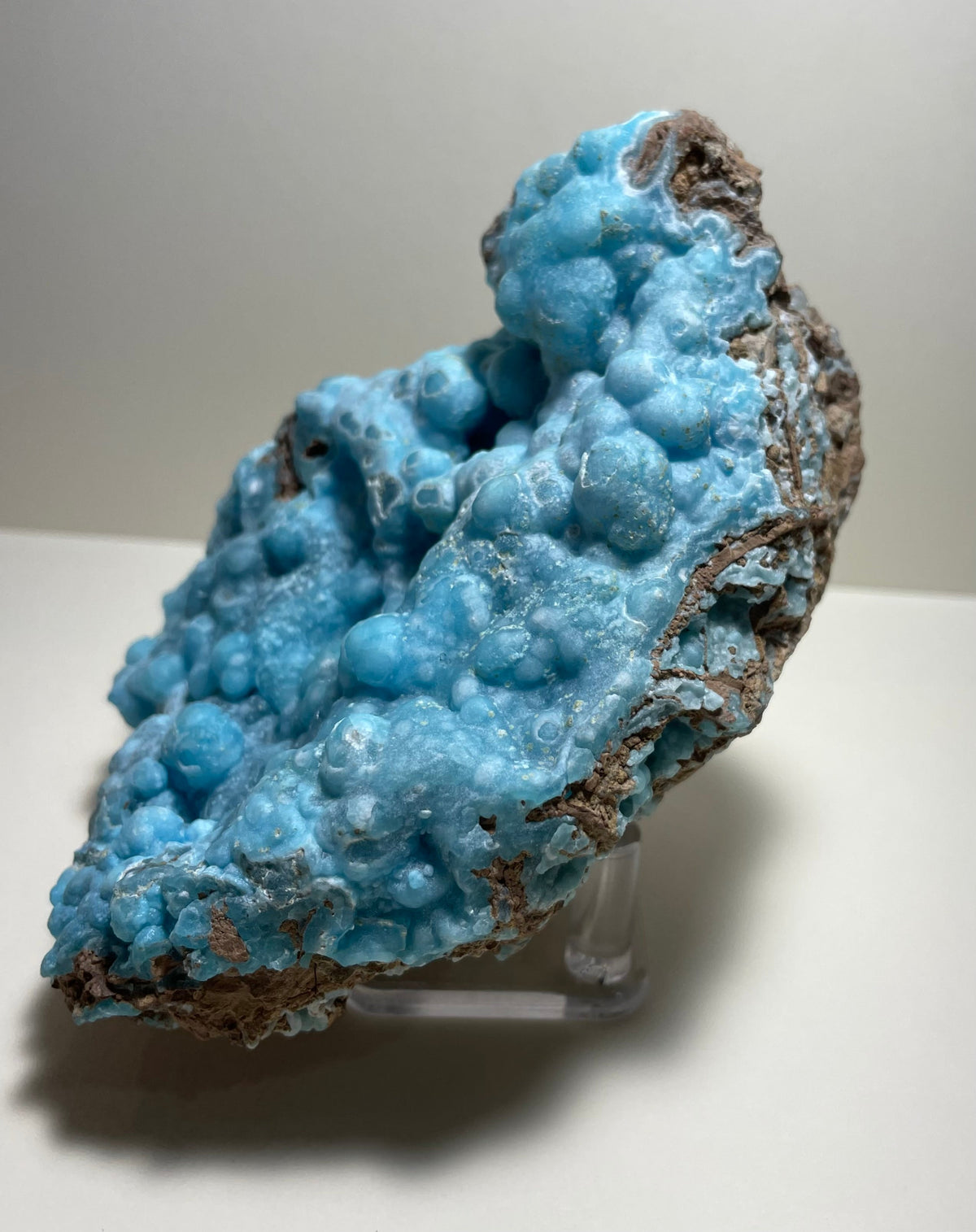 Botryoidal blue hemimorphite specimen with rounded, bubbly surface texture and brown mineral matrix, displayed on a clear plastic stand against a light background.